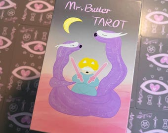 May include: A tarot card deck box with the words "Mr. Butter Tarot" and a whimsical illustration. The artwork features two purple serpent-like figures embracing a central figure holding pink swords, set against a gradient background with a crescent moon.