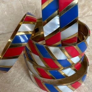 Puede incluir: Una pila de seis pulseras de brazalete con un patrón de rayas rojas, blancas y azules. Las pulseras están hechas de latón y tienen un acabado dorado. Las rayas son diagonales y las pulseras están apiladas una encima de la otra.