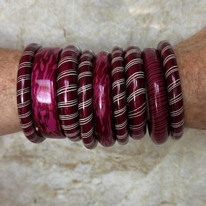 Op de afbeelding: Een verzameling bordeauxrode armbanden met witte streepaccenten. Sommige armbanden hebben een effen bordeauxrode kleur, terwijl andere een patroon hebben. De armbanden zijn op een pols gestapeld en tonen hun verschillende stijlen en texturen.