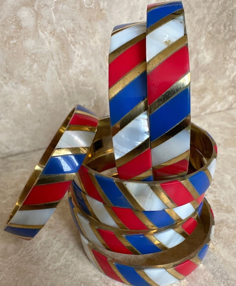 Puede incluir: Una pila de seis pulseras de brazalete con un patr&oacute;n de rayas rojas, blancas y azules. Las pulseras est&aacute;n hechas de lat&oacute;n y tienen un acabado dorado. Las rayas son diagonales y las pulseras est&aacute;n apiladas una encima de la otra.