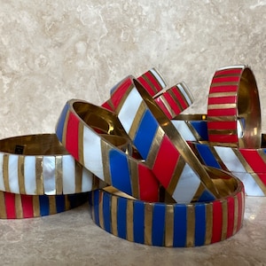 Puede incluir: Una pila de pulseras coloridas con rayas rojas, blancas y azules. Las pulseras son de lat&oacute;n y tienen un acabado brillante.
