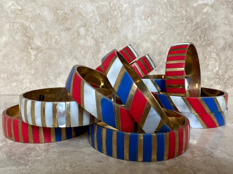 Puede incluir: Una pila de pulseras coloridas con rayas rojas, blancas y azules. Las pulseras son de lat&oacute;n y tienen un acabado brillante.
