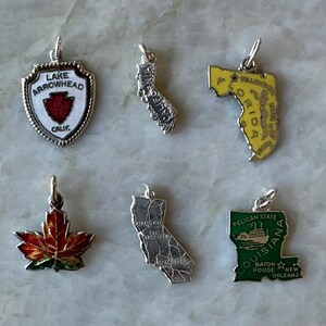 May include: A collection of silver-toned and enameled charms. The charms depict a shield with "Lake Arrowhead Calif", a maple leaf, two California state outlines, a Florida outline, and a Louisiana outline with "Pelican State".