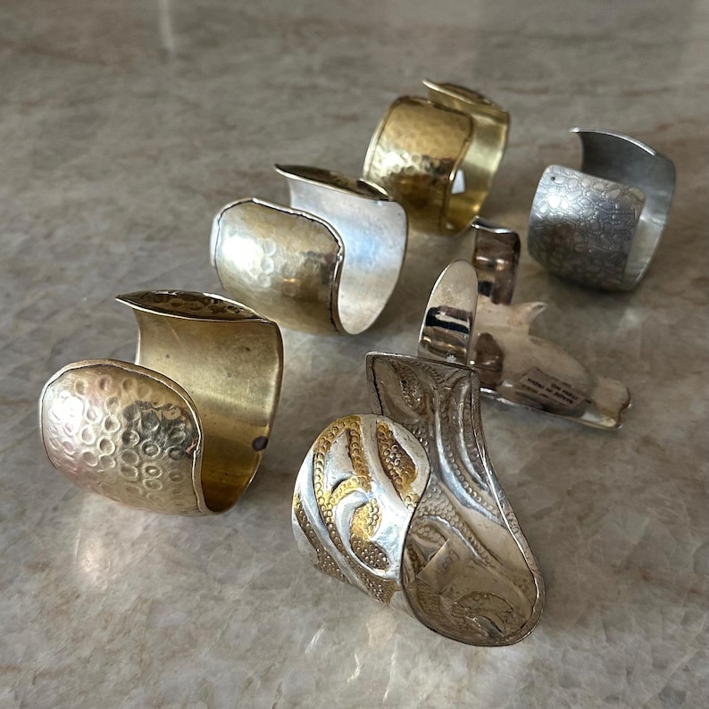 May include: A collection of handcrafted metal rings in various designs. The rings feature textures like hammered and engraved patterns, and are in shades of gold and silver. Some rings have a wide band, while others have a more open design.