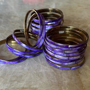 May include: Stack of gold-toned bangles with a mosaic of purple and white rectangular inlays. Several bangles are scattered around the stack, showcasing the design. The bangles are arranged on a light-colored surface.