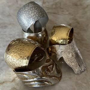 May include: An assortment of metal napkin rings in silver and gold tones. The rings feature various textures, including hammered, patterned, and embossed designs. One ring is shaped like a horse head. The napkin rings are stacked on a light-colored surface.