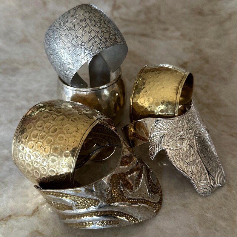 May include: An assortment of metal napkin rings in silver and gold tones. The rings feature various textures, including hammered, patterned, and embossed designs. One ring is shaped like a horse head. The napkin rings are stacked on a light-colored surface.