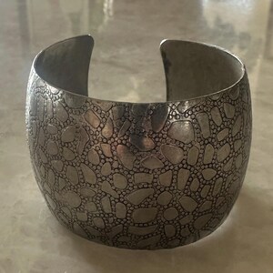 May include: A wide, silver-toned cuff bracelet with a textured surface. The bracelet features an all-over pattern of irregular shapes and small, raised dots. The open design allows for easy wear.