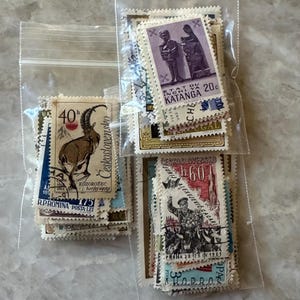 Op de afbeelding: Een verzameling vintage postzegels in doorzichtige plastic zakken. De postzegels tonen diverse ontwerpen, waaronder een steenbok, figuren en portretten. Sommige postzegels hebben tekst in meerdere talen, met waarden zoals 40h en 20c.