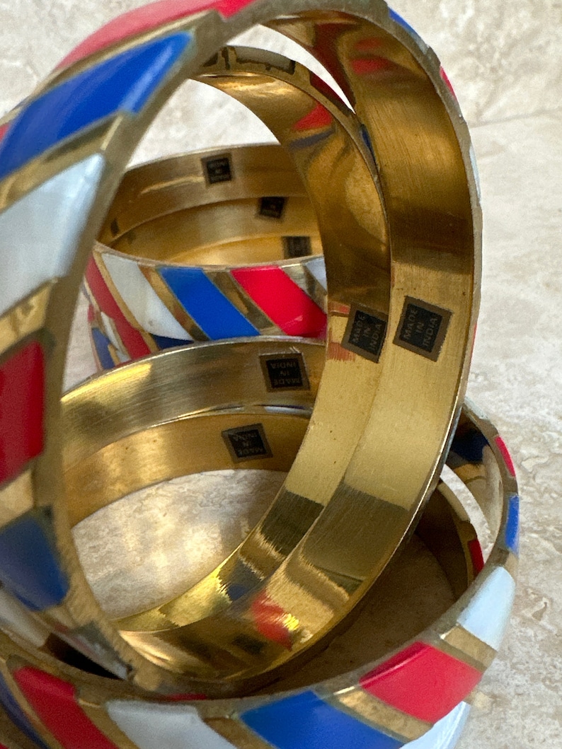 Puede incluir: Conjunto de cuatro pulseras de oro con rayas de esmalte rojo, blanco y azul. Cada pulsera tiene las palabras "Made in India" estampadas en el interior.