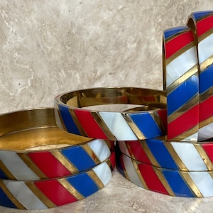 Puede incluir: Conjunto de seis pulseras de oro con esmalte rayado rojo, blanco y azul. Las rayas son diagonales y las pulseras est&aacute;n apiladas unas sobre otras.
