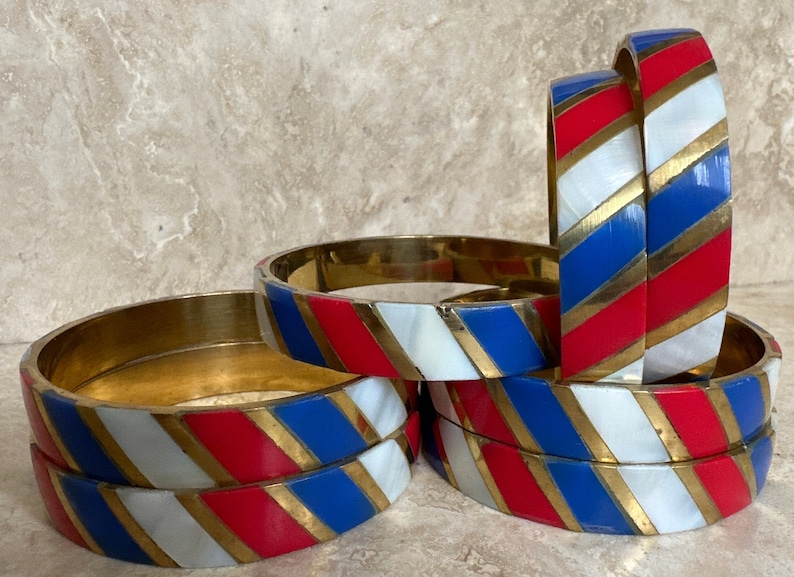 Puede incluir: Conjunto de seis pulseras de oro con esmalte rayado rojo, blanco y azul. Las rayas son diagonales y las pulseras est&aacute;n apiladas unas sobre otras.