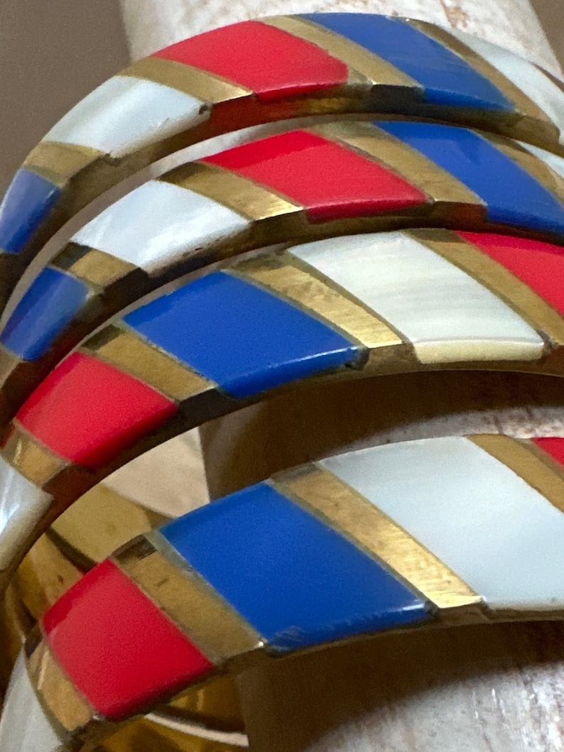 Puede incluir: Conjunto de cuatro brazaletes dorados con esmalte rayado rojo, blanco y azul. Las rayas son diagonales y los brazaletes est&aacute;n apilados uno encima del otro.