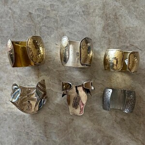 May include: Collection of six metal cuff bracelets in various designs. Includes gold-toned hammered metal, silver with etched patterns, and a silver cuff with a floral design. The bracelets are displayed on a light-colored surface.
