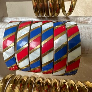 Puede incluir: Un conjunto de seis pulseras coloridas con rayas rojas, blancas, azules y doradas. Las pulseras son de metal y tienen un acabado brillante.