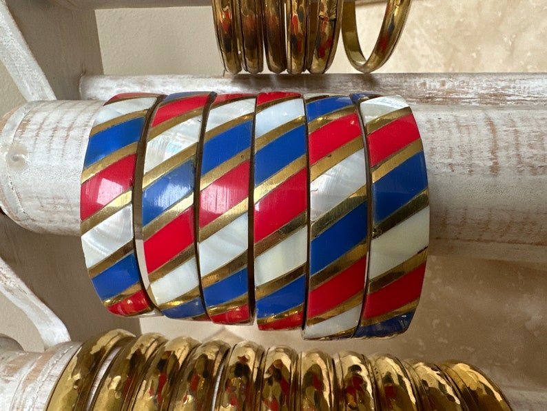 Puede incluir: Un conjunto de seis pulseras coloridas con rayas rojas, blancas, azules y doradas. Las pulseras son de metal y tienen un acabado brillante.