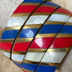 Puede incluir: Conjunto de cuatro pulseras de brazalete con un patr&oacute;n a rayas en rojo, blanco, azul y dorado. Las pulseras est&aacute;n hechas de un material duro y brillante.