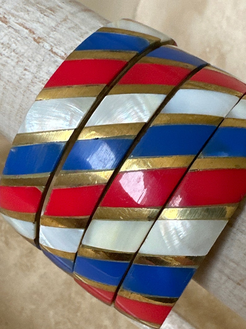 Puede incluir: Conjunto de cuatro pulseras de brazalete con un patr&oacute;n a rayas en rojo, blanco, azul y dorado. Las pulseras est&aacute;n hechas de un material duro y brillante.