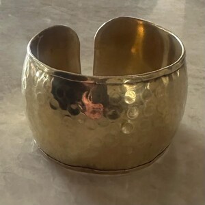 May include: A wide, gold-colored cuff bracelet with a hammered texture. The open design allows for easy wear. The bracelet has a polished finish and a slightly rounded shape, suitable for various wrist sizes.