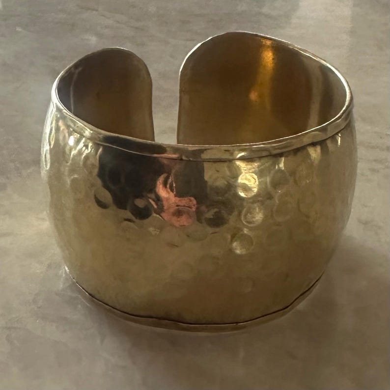 May include: A wide, gold-colored cuff bracelet with a hammered texture. The open design allows for easy wear. The bracelet has a polished finish and a slightly rounded shape, suitable for various wrist sizes.