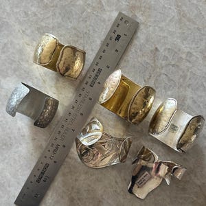 May include: An assortment of silver and gold-toned metal cuff bracelets. Some bracelets have a hammered texture, while others feature intricate patterns. A ruler is placed next to the bracelets.