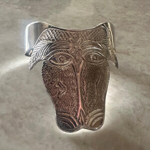May include: A silver-toned cuff bracelet featuring a stylized face design. The face has detailed eyes, a nose, and a textured surface. The bracelet is approximately 2 inches wide.