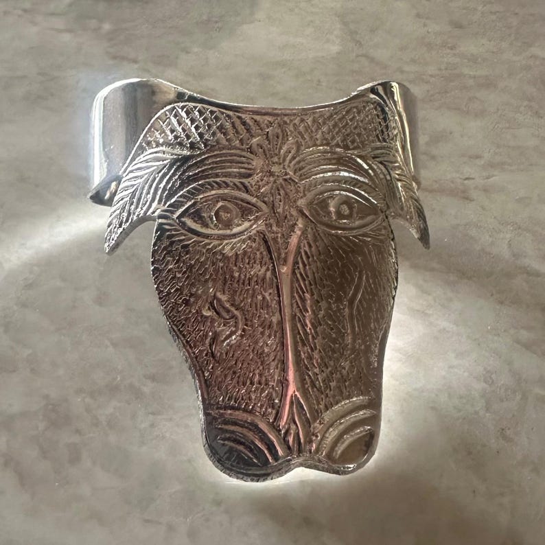May include: A silver-toned cuff bracelet featuring a stylized face design. The face has detailed eyes, a nose, and a textured surface. The bracelet is approximately 2 inches wide.