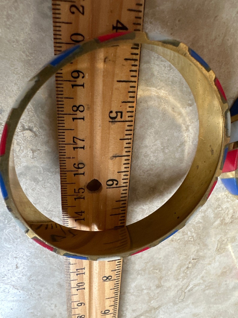 Puede incluir: Un brazalete de lat&oacute;n con un patr&oacute;n geom&eacute;trico rojo, blanco y azul. El brazalete tiene un di&aacute;metro de aproximadamente 5 cm.