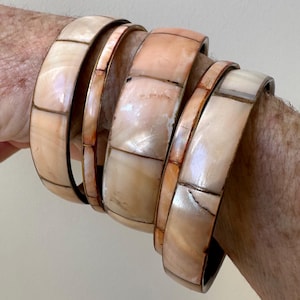 Może przedstawiać: Zestaw czterech bransoletek z muszli i mosiądzu. Bransoletki są wykonane z jasnoróżowej muszli z geometrycznym wzorem. Mosiądz ma ciepły, czerwonobrązowy kolor.