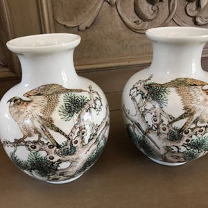 Op de afbeelding: Twee witte porseleinen vazen met een vogel- en boomontwerp. De vazen zijn versierd met een bruine en witte vogel die op een tak zit met groene bladeren.