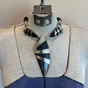 May include: A statement necklace with a large, geometric pendant. The pendant features black and silver stripes and a silver teardrop shape. The necklace has a black cord and silver bead accents, displayed on a mannequin.