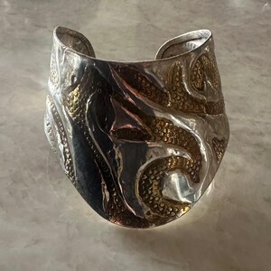 May include: Wide silver cuff bracelet with gold-toned accents. The bracelet has a textured surface with an embossed design. The cuff is wide and has a curved shape, with a polished finish. The gold accents are in a repeating pattern.