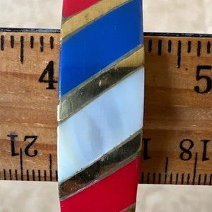 Puede incluir: Un brazalete de rayas rojas, blancas y azules con detalles dorados. El brazalete tiene un di&aacute;metro de aproximadamente 10 cm.