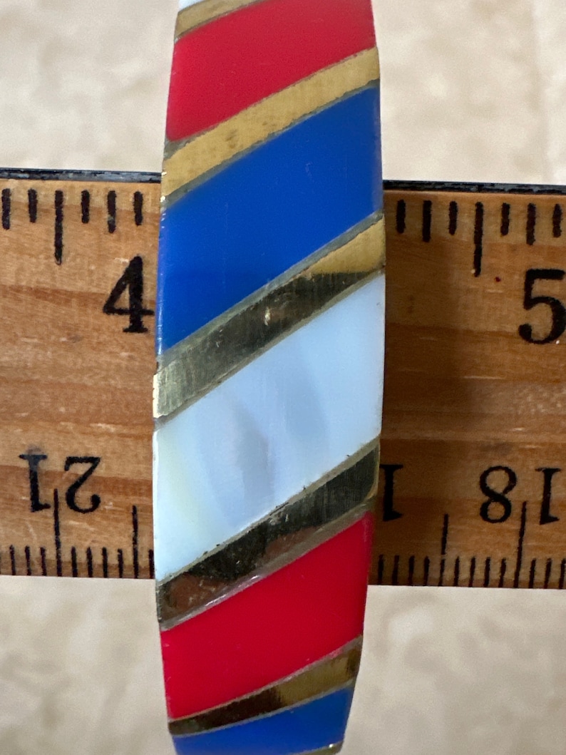 Puede incluir: Un brazalete de rayas rojas, blancas y azules con detalles dorados. El brazalete tiene un di&aacute;metro de aproximadamente 10 cm.