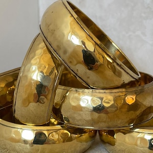 Pode incluir: Um conjunto de pulseiras douradas com textura martelada. As pulseiras são grossas e brilhantes, refletindo a luz. As pulseiras estão empilhadas, criando um efeito em camadas. A estética geral é vintage e elegante. Cada pulseira tem cerca de 6,5 cm de diâmetro.
