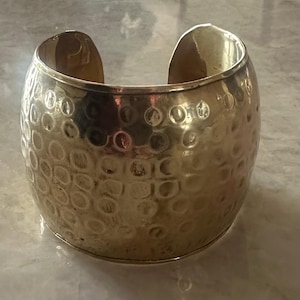 May include: A wide, gold-colored cuff bracelet with a textured, hammered surface. The bracelet has an open design and is made of a shiny metal. The cuff is a statement piece of jewelry.