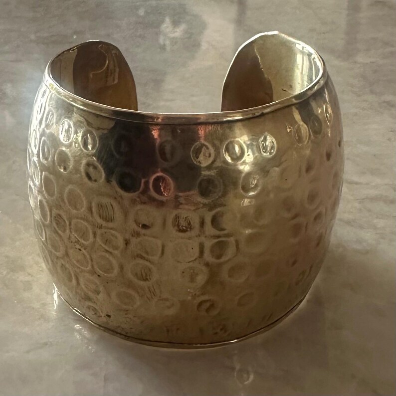 May include: A wide, gold-colored cuff bracelet with a textured, hammered surface. The bracelet has an open design and is made of a shiny metal. The cuff is a statement piece of jewelry.