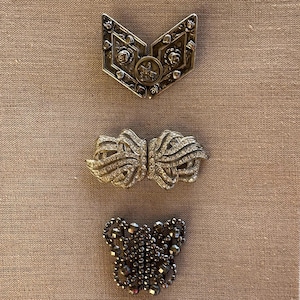 Puede incluir: Tres broches vintage de diferentes estilos. La broche superior es un diseño de corazón plateado con detalles florales. La broche del medio es un diseño plateado y de diamantes de imitación con un patrón de remolino. La broche inferior es un diseño negro y plateado con un patrón geométrico.