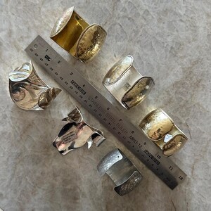 May include: An assortment of silver and gold-toned metal cuff bracelets. Some have hammered textures, while others feature intricate patterns. A ruler is placed next to the bracelets for scale.