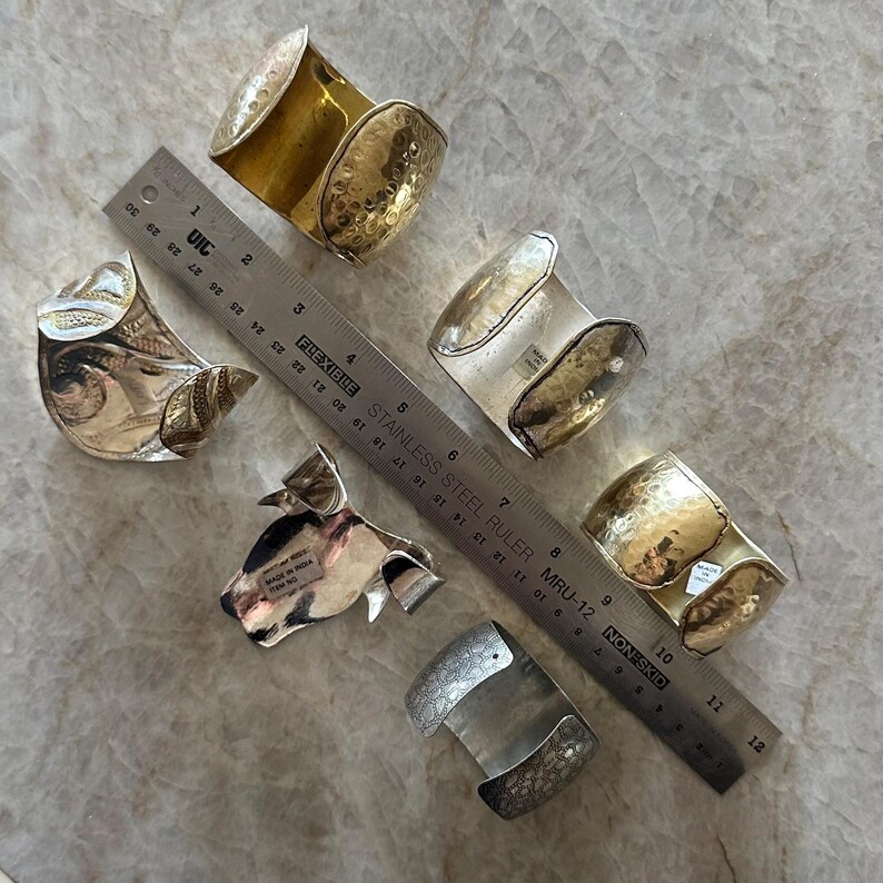 May include: An assortment of silver and gold-toned metal cuff bracelets. Some have hammered textures, while others feature intricate patterns. A ruler is placed next to the bracelets for scale.