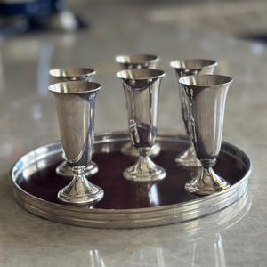 Puede incluir: Seis copas plateadas con bordes acampanados y bases de pedestal dispuestas sobre una bandeja redonda marrón oscuro con borde plateado. Las copas son reflectantes y la bandeja descansa sobre una superficie clara.