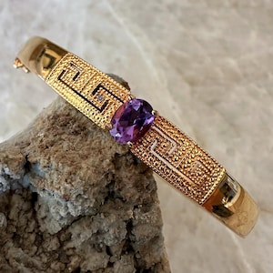 Op de afbeelding: Een goudkleurige scharnierende armband met een centrale ovaal geslepen paarse edelsteen. De band is versierd met een herhalend geometrisch Grieks sleutelpatroon en een textuurafwerking, met een kleine heldere steen nabij de centrale zetting.