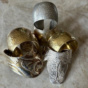 May include: An assortment of metal bracelets and a decorative face-shaped object. The bracelets include silver and gold-toned pieces with textured and patterned surfaces. The face-shaped object is silver with detailed facial features.