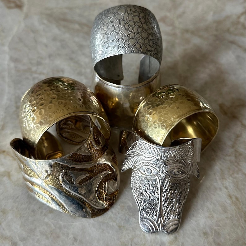 May include: An assortment of metal bracelets and a decorative face-shaped object. The bracelets include silver and gold-toned pieces with textured and patterned surfaces. The face-shaped object is silver with detailed facial features.