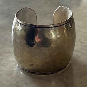 May include: A wide, silver-toned cuff bracelet with a hammered texture. The open-ended design allows for easy wear. The bracelet has a slightly oval shape and a polished finish, reflecting light.