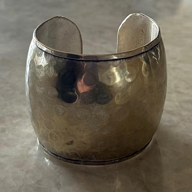 May include: A wide, silver-toned cuff bracelet with a hammered texture. The open-ended design allows for easy wear. The bracelet has a slightly oval shape and a polished finish, reflecting light.