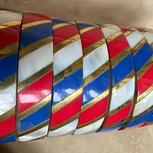Puede incluir: Un conjunto de siete pulseras coloridas con un patr&oacute;n a rayas en rojo, blanco, azul y dorado. Las pulseras est&aacute;n hechas de un material brillante y est&aacute;n apiladas unas sobre otras.