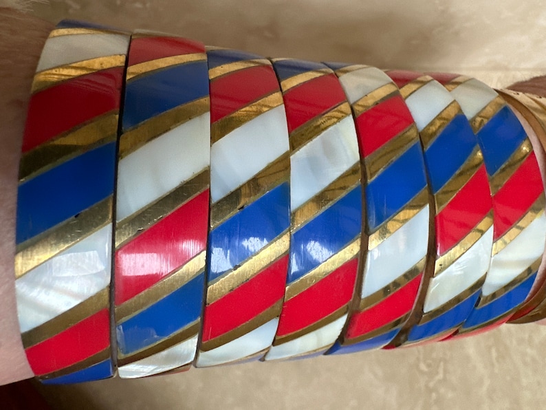 Puede incluir: Un conjunto de siete pulseras coloridas con un patr&oacute;n a rayas en rojo, blanco, azul y dorado. Las pulseras est&aacute;n hechas de un material brillante y est&aacute;n apiladas unas sobre otras.