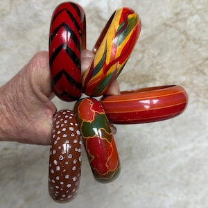 以下が含まれることがあります： さまざまなデザインと色の木製バングル5つのコレクション。1つのバングルは黒のシェブロンストライプの赤、もう1つはカラフルな抽象模様、3つ目は白い水玉模様の茶色です。他の2つはオレンジ色のストライプの赤と地図デザインの赤です。