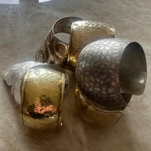 May include: An assortment of silver and gold-toned metal cuff bracelets. The bracelets feature various textures, including hammered and patterned designs. One bracelet has a detailed floral design, while others have a smooth, polished finish.